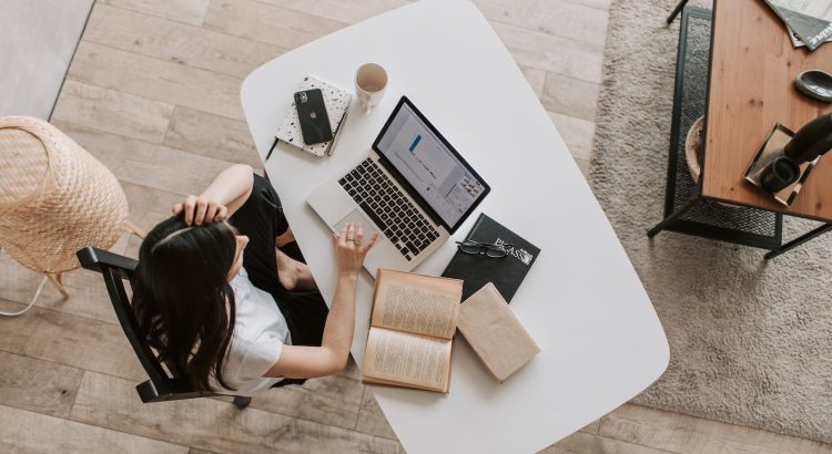 From above of young woman with long dark hair in casual clothes working at table and browsing netbook while sitting in modern workplace and touching hair